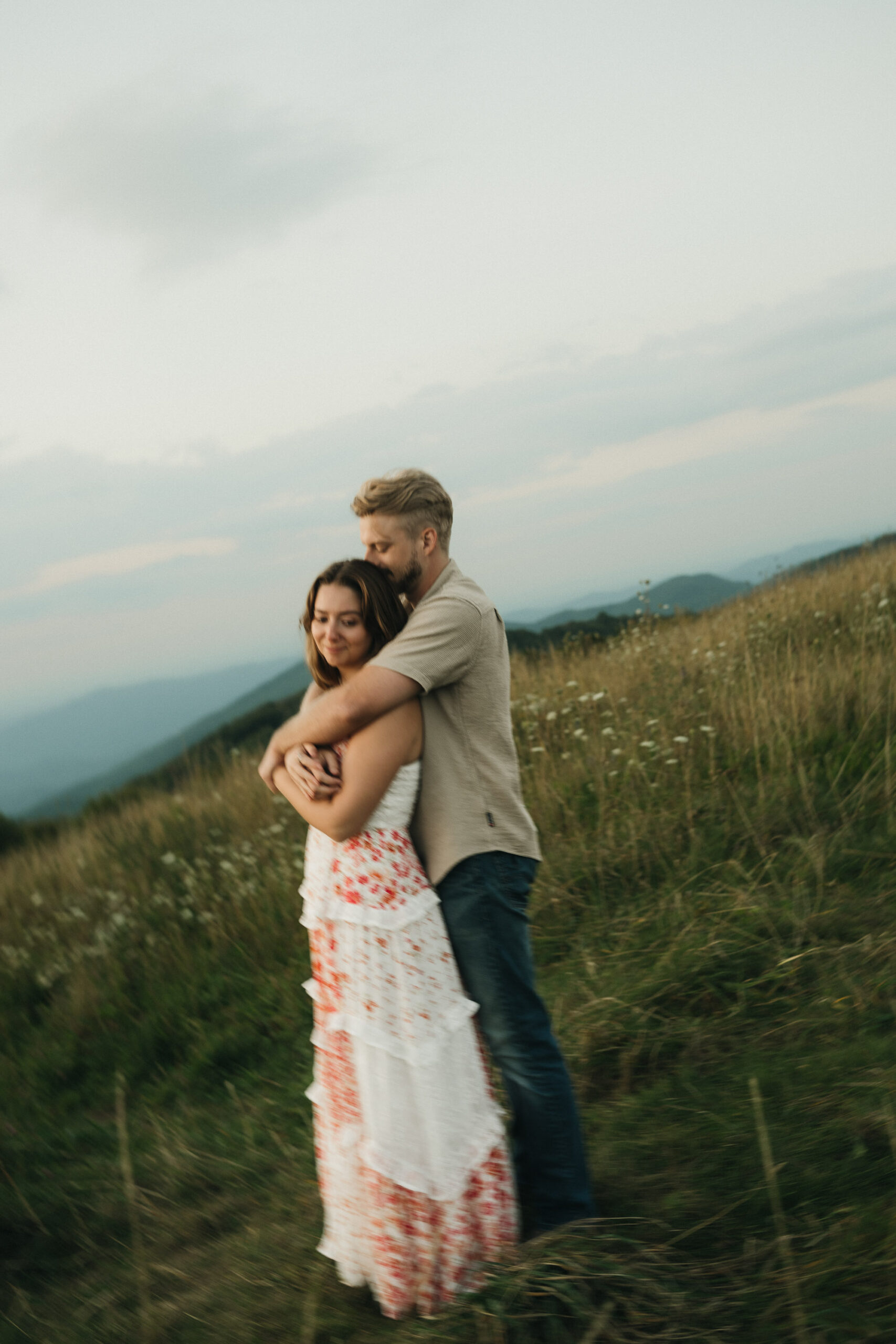 A man stands in a grassy field hugging a woman from behind. The woman wears a white and pink dress, and the man wears a beige shirt and jeans. The background features rolling hills under a cloudy sky. Asheville wedding photographer