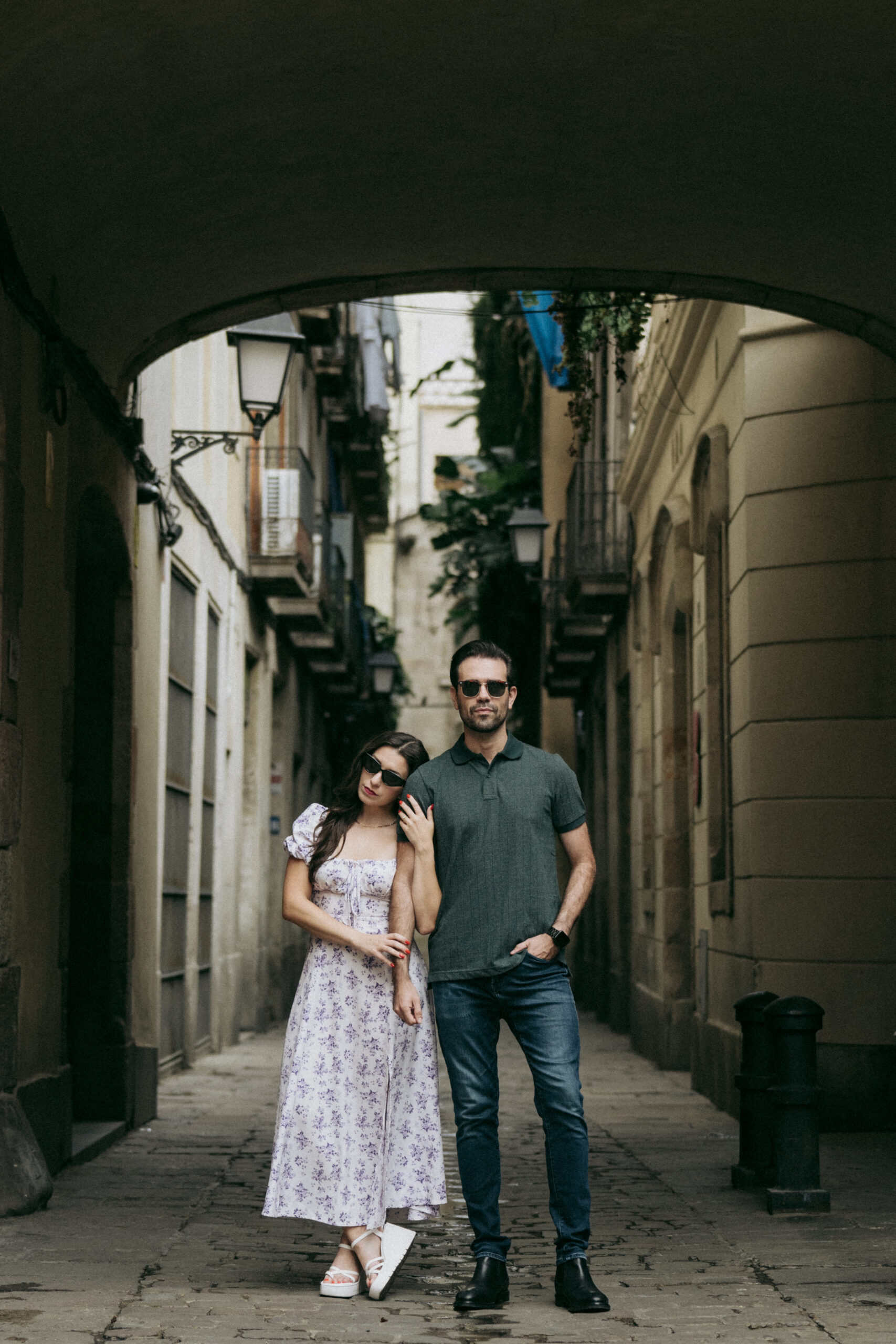 A woman in a floral dress leans on a man in a green shirt and sunglasses as they stand together in a narrow, shaded European street with stone buildings and balconies. Asheville wedding photographer