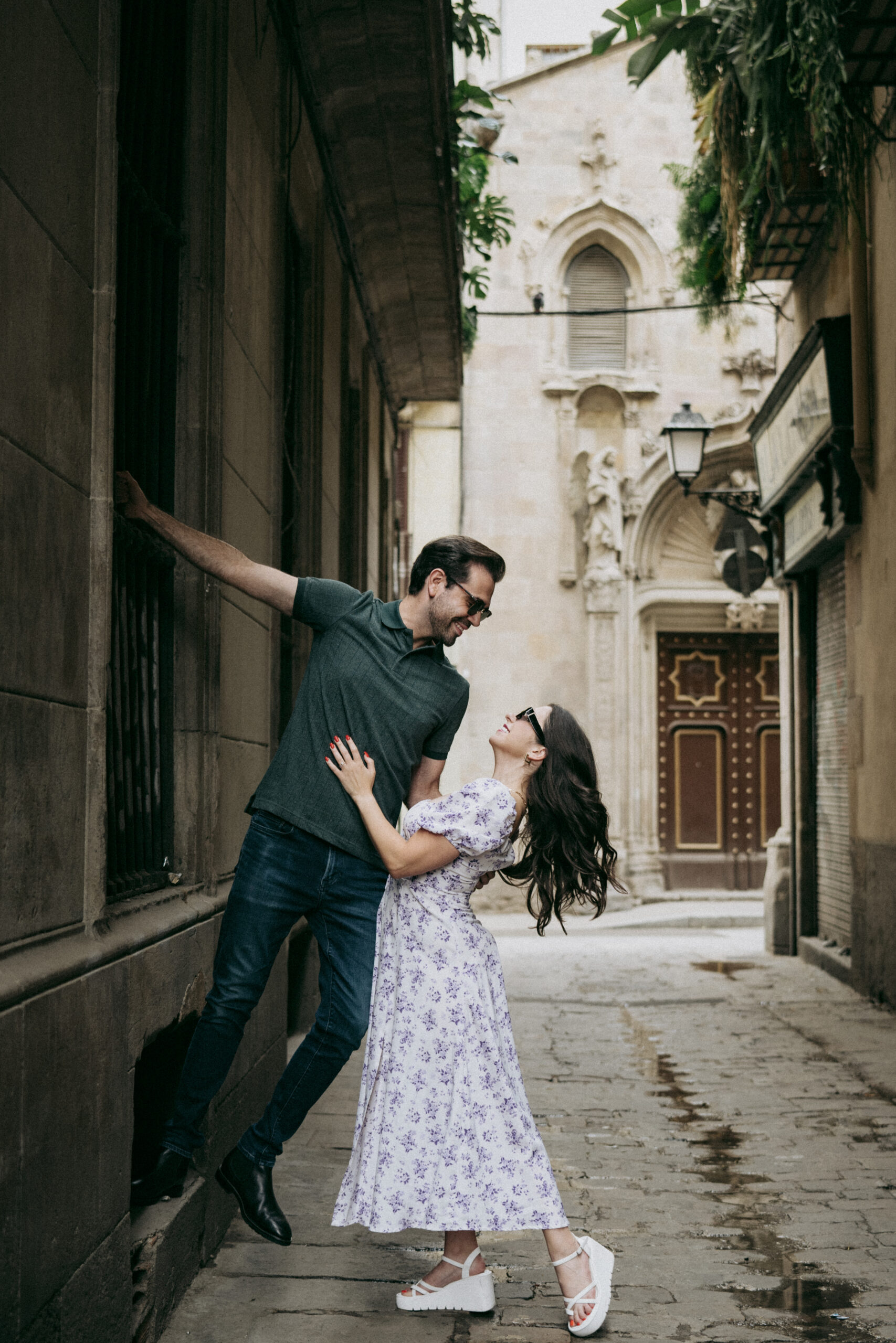 A man playfully leans against a wall while a woman in a floral dress holds his arm and smiles at him. They are in a charming, narrow street with historic stone buildings in the background. Asheville wedding photographer