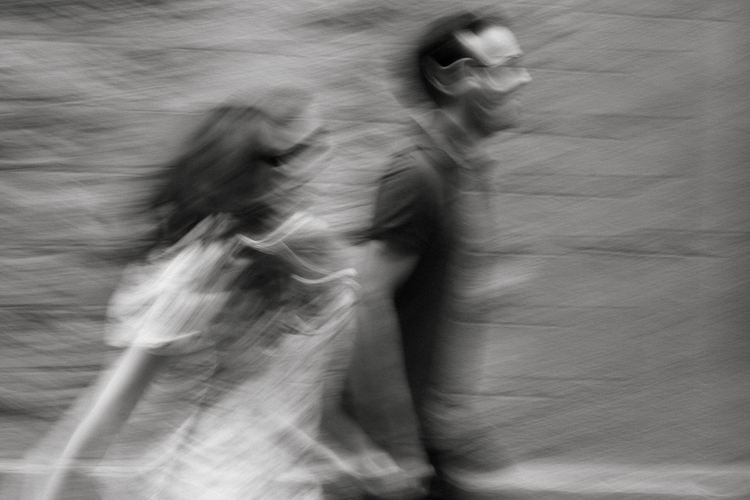 A blurry black-and-white photo of two people walking side by side, likely a man and a woman, with indistinct features and motion blur suggesting movement. The background appears to be a textured wall. Asheville wedding photographer