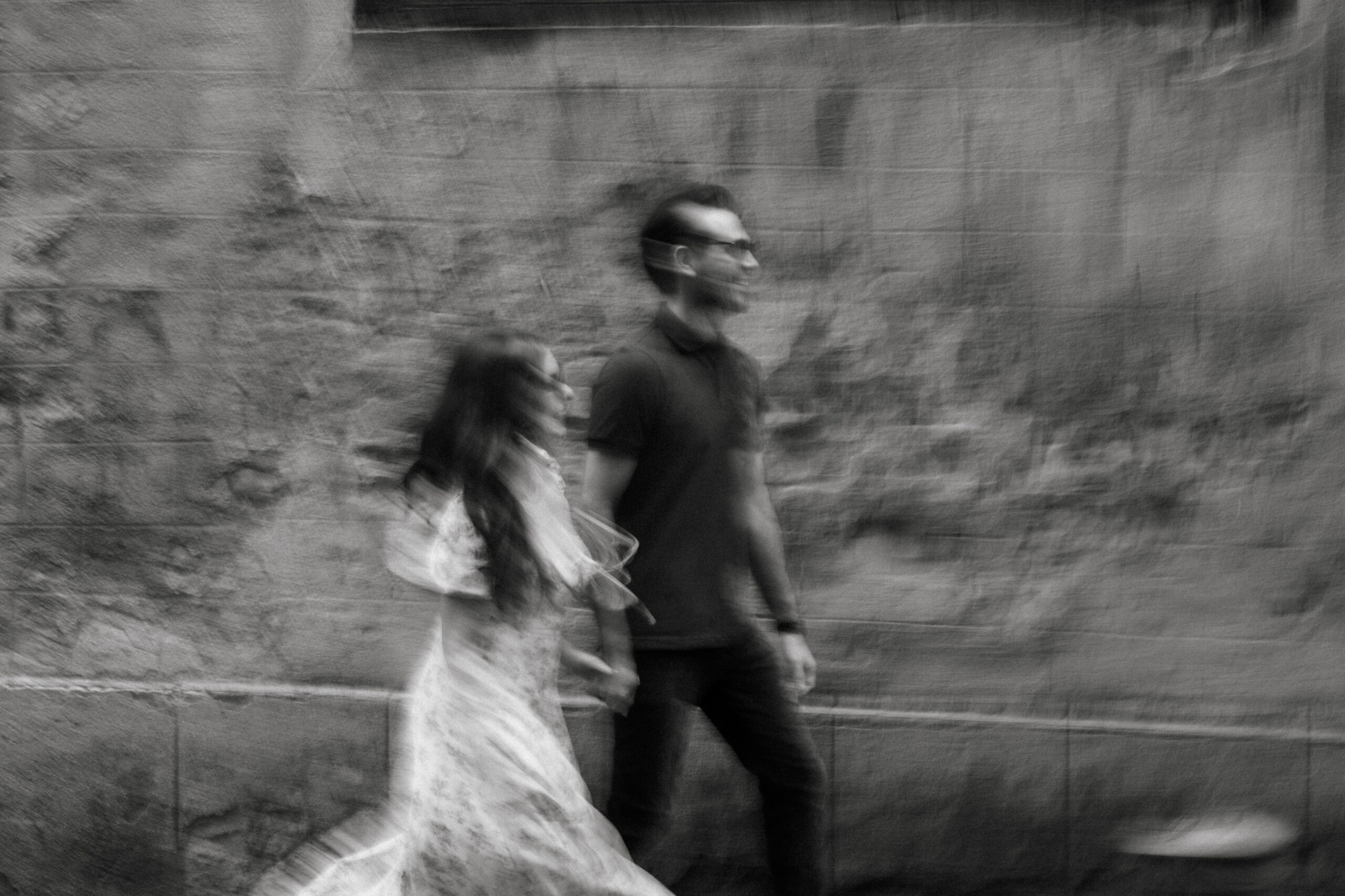 A black and white, blurred photo of a man and woman walking side by side along a textured wall. The woman wears a long dress; the man wears a polo shirt and glasses. Both appear to be moving. Asheville wedding photographer