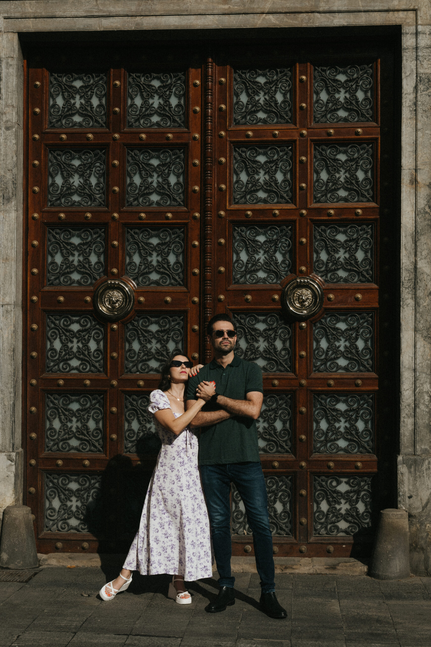 A woman in a floral dress and sunglasses stands arm-in-arm with a man in dark clothes and sunglasses, posing in front of a large ornate wooden door with intricate metalwork and lion head knockers. Asheville wedding photographer