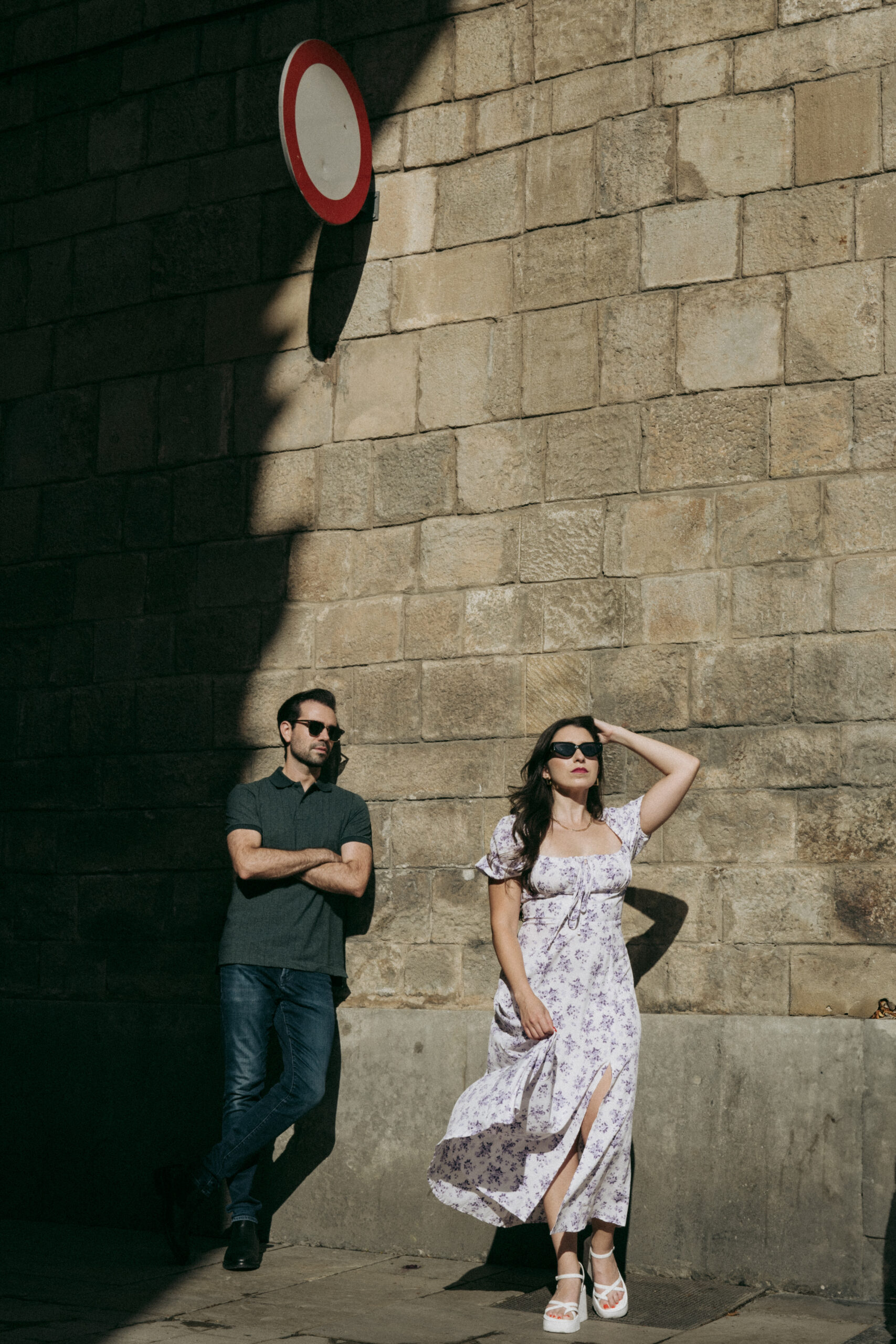 A man in dark clothing stands with arms crossed next to a woman in a white floral dress and sunglasses, posing against a sunlit brick wall beneath a red and white traffic sign. Asheville wedding photographer