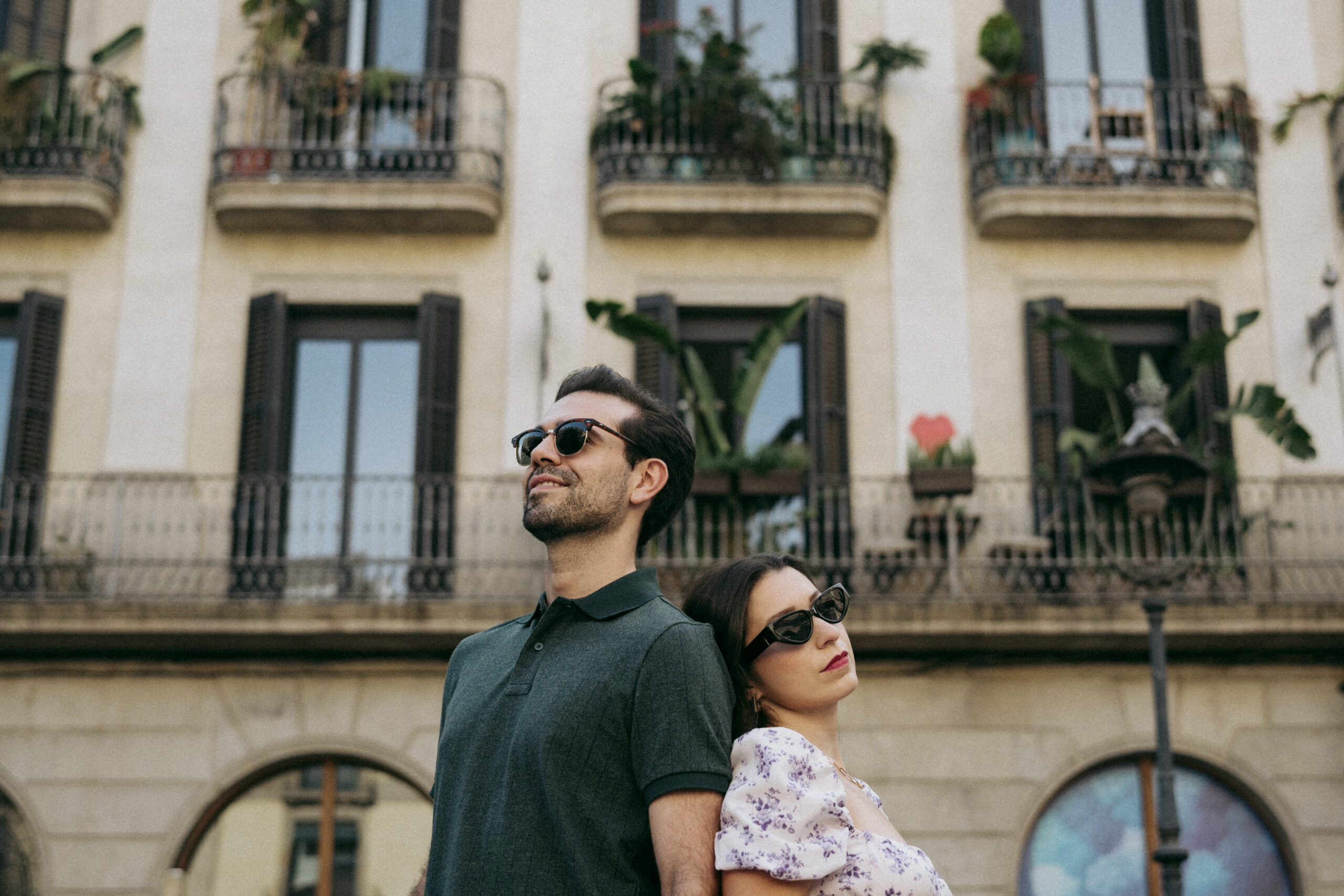 A man and woman wearing sunglasses stand back to back, looking in opposite directions, in front of a building with balconies and plants. Asheville wedding photographer