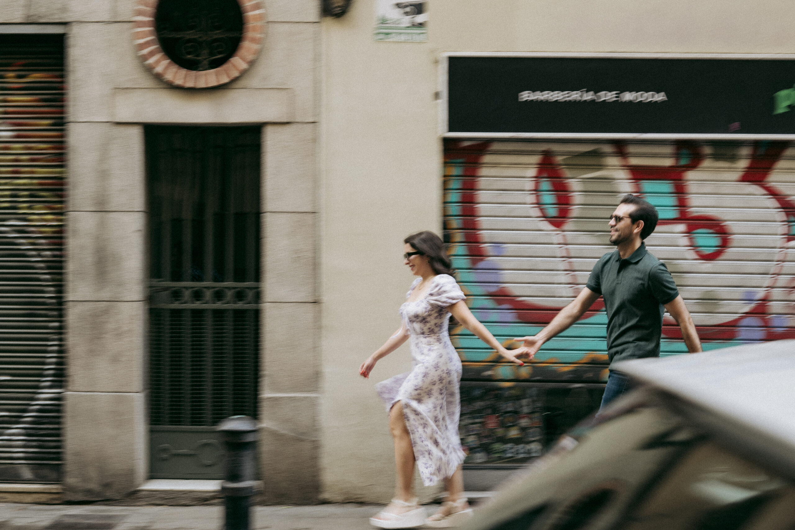 A couple walks hand in hand down a city street, smiling. The woman wears a floral dress and sunglasses, and the man wears a dark shirt and pants. Graffiti and shop signs are visible on the buildings behind them. Asheville wedding photographer