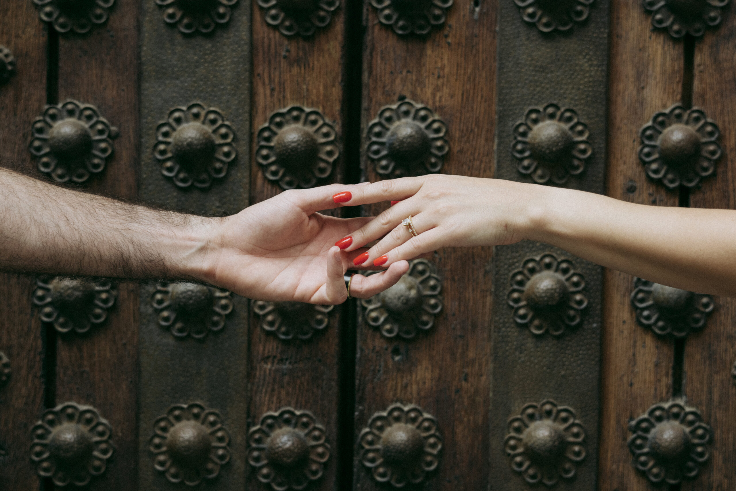 Two hands reaching toward each other in front of an ornate wooden door with decorative metal studs; one hand has red nail polish and a ring, the other is bare. Asheville wedding photographer