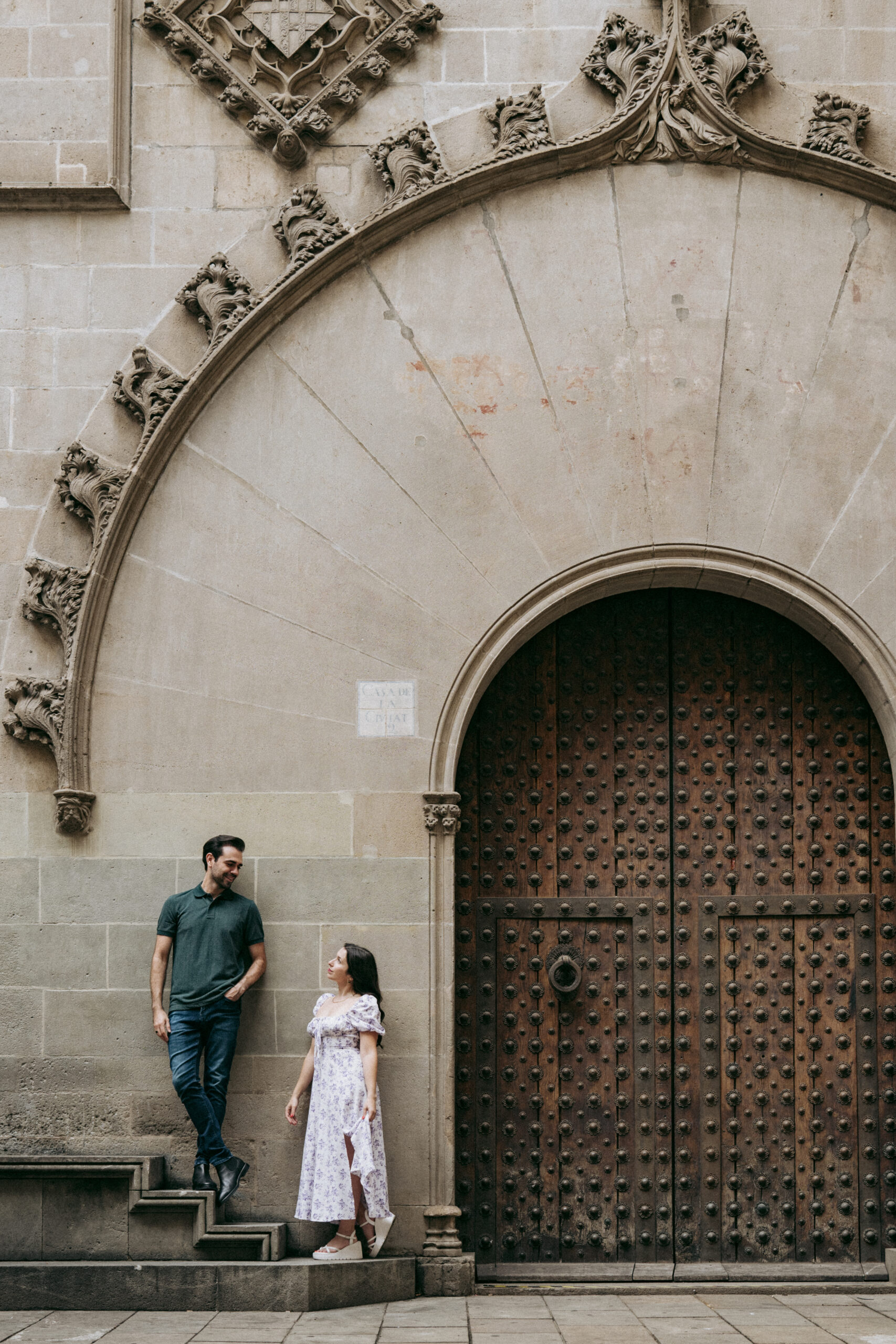 A man and woman stand beside a large ornate wooden door set in a stone wall with intricate Gothic architectural details. The man stands on steps, looking down at the woman, who stands on the ground, looking up at him. Asheville wedding photographer