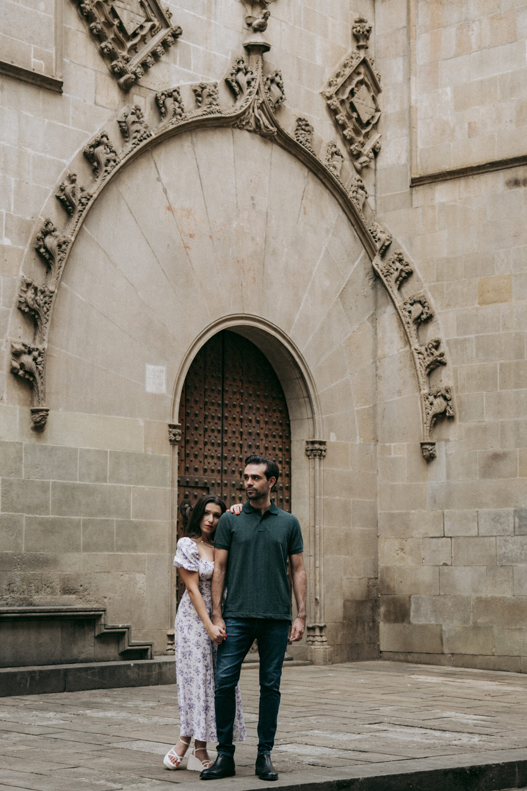 A couple stands close together, holding hands in front of an ornate, arched doorway set in a stone building. The woman wears a light floral dress and sandals, and the man wears a green polo shirt and jeans. Asheville wedding photographer