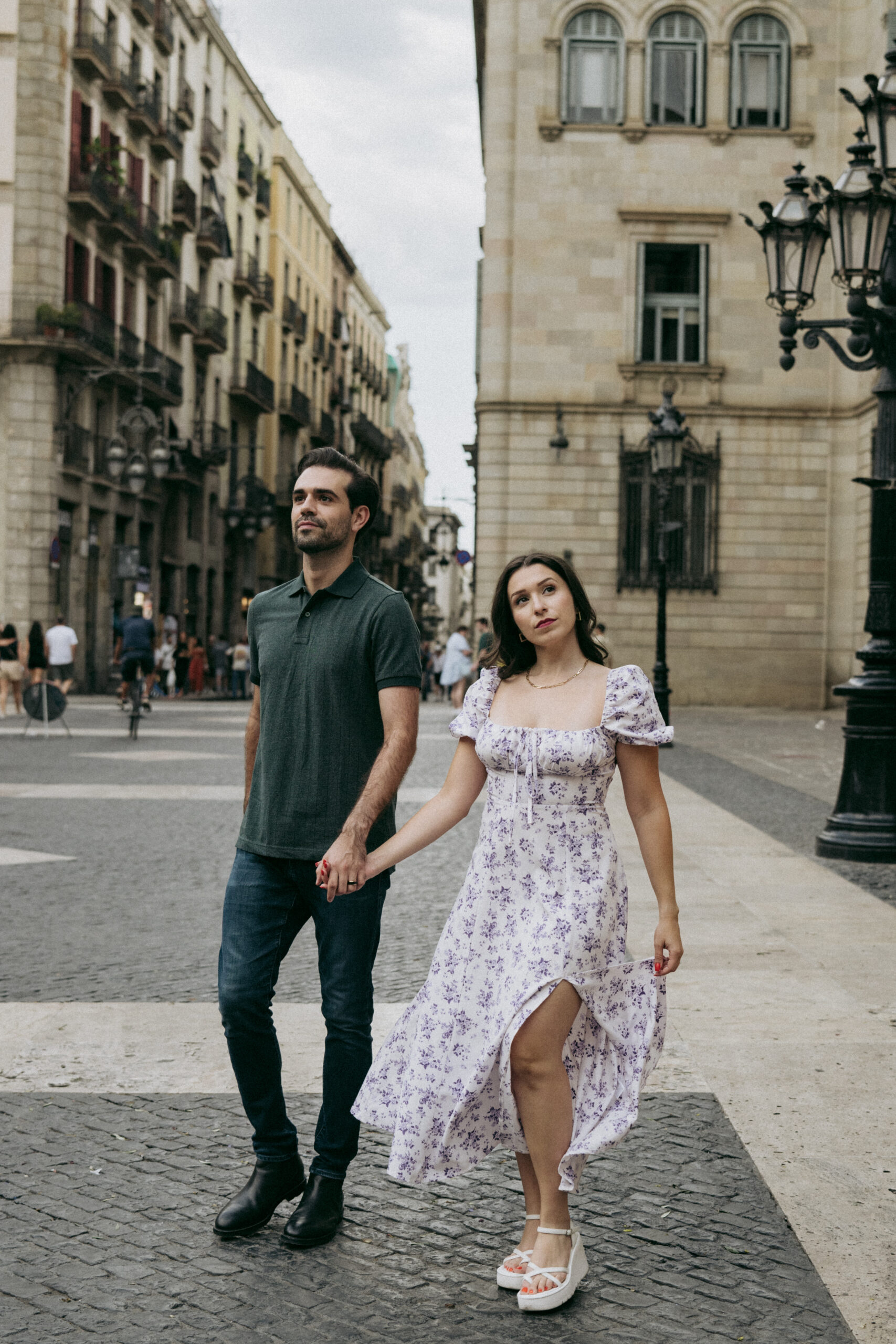 A man and a woman hold hands while walking on a city street. The woman wears a white and purple floral dress, and the man wears a dark shirt and jeans. They pass historic buildings and old-fashioned street lamps. Asheville wedding photographer