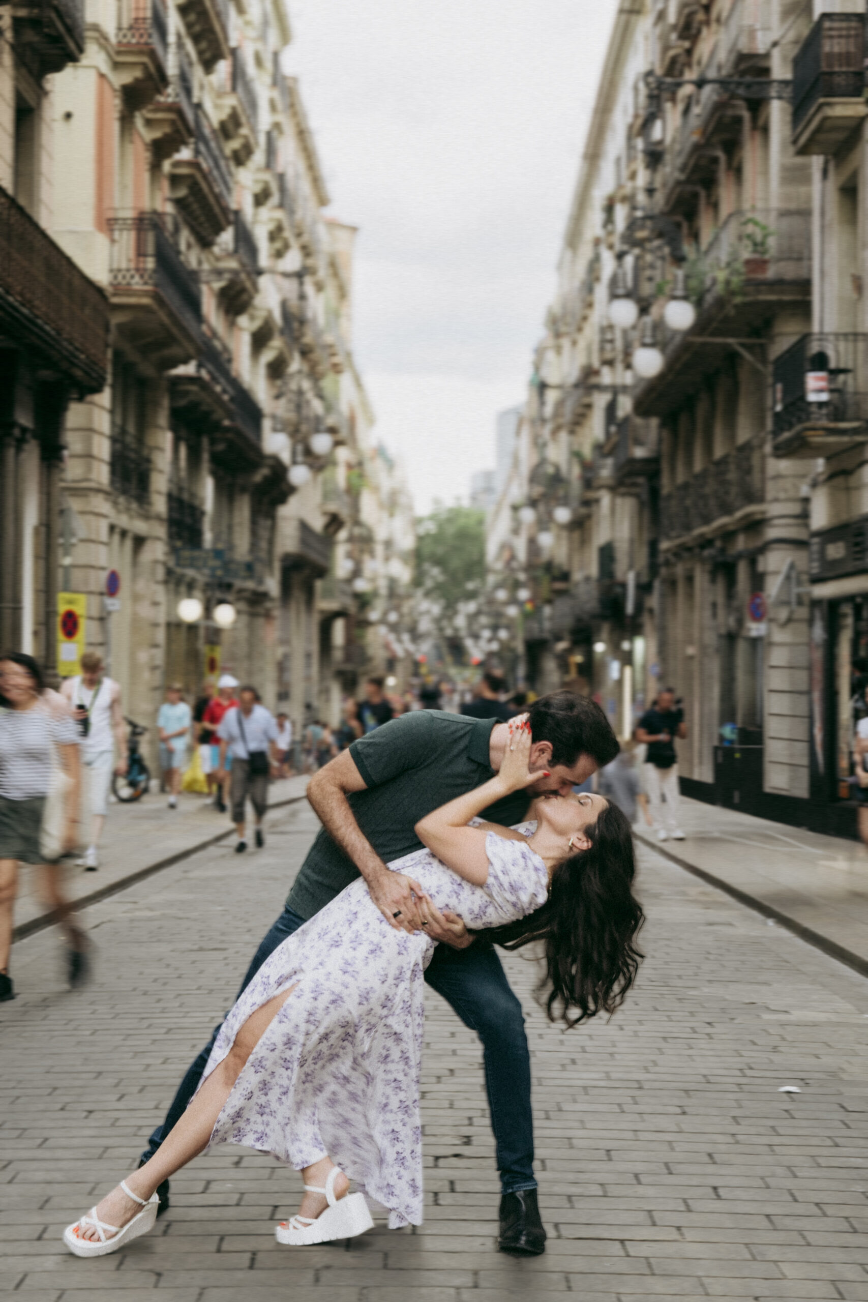 A couple dances in the middle of a lively, European city street, with the man dipping the woman and both sharing a kiss. People walk by around them, blurred in motion. Tall historic buildings line both sides of the street. Asheville wedding photographer