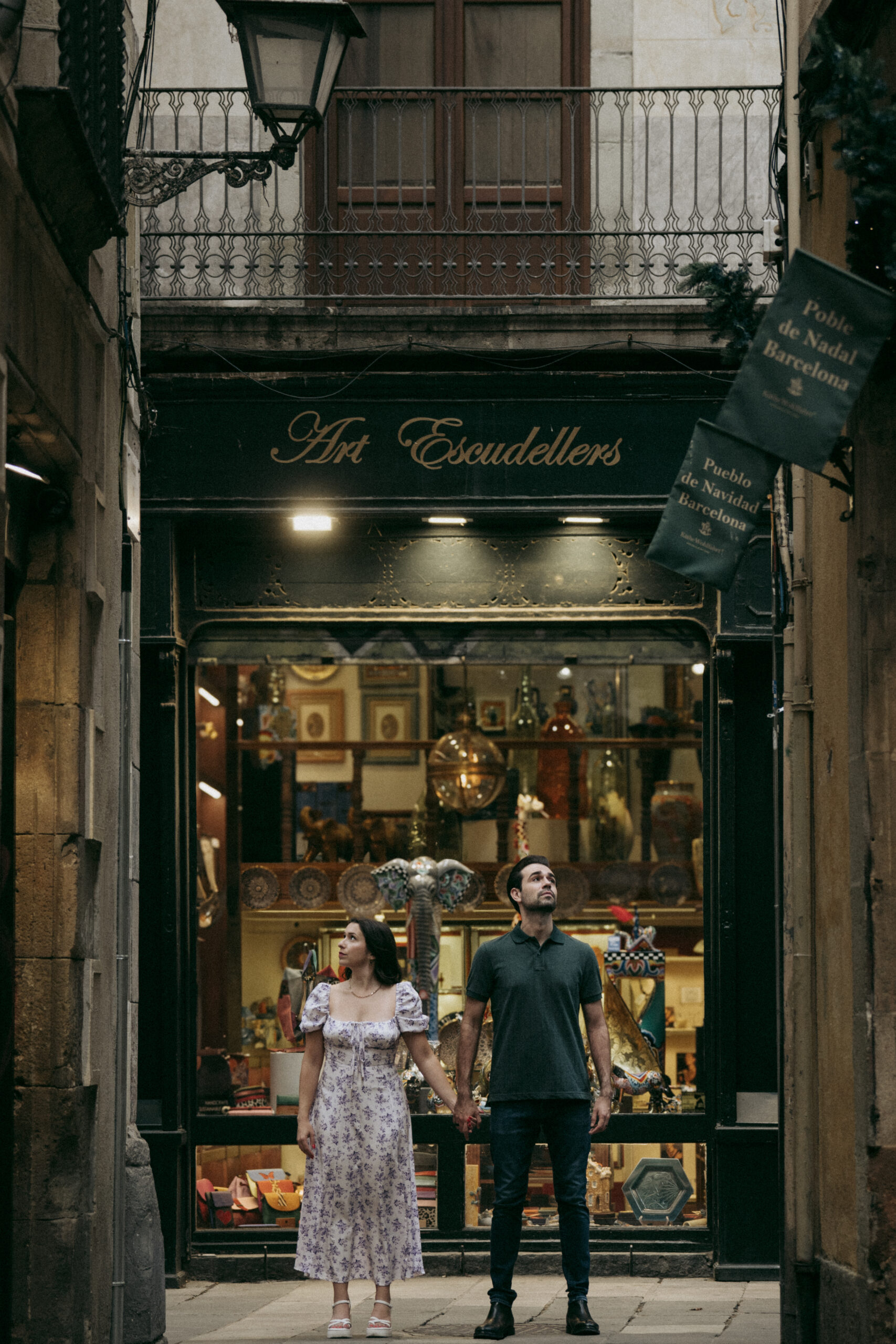 A couple stands holding hands in front of a shop called Art Escudellers on a narrow street, looking up. The shop window displays various colorful items inside. The scene has a vintage, urban atmosphere. Asheville wedding photographer