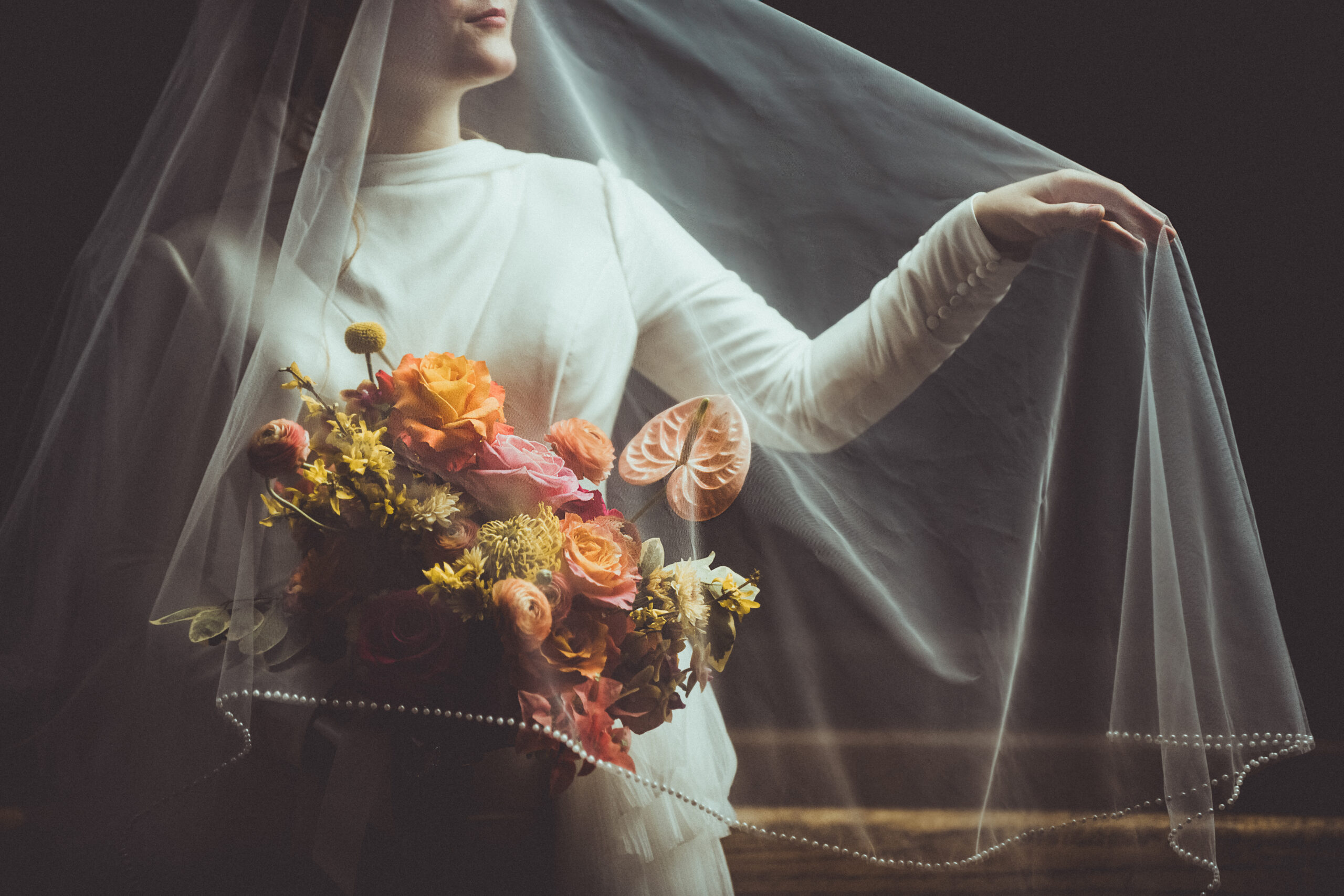 Fine art wedding photography in Asheville – a bride in an elegant long-sleeve gown holds a romantic bouquet of peach, pink, and golden florals beneath a delicate pearl-trimmed veil. Captured in soft, moody lighting, this timeless bridal portrait embodies luxury and sophistication. Asheville wedding photographer specializing in high-end, artistic weddings.
Bride holding colorful bouquet under veil, fine art wedding photography in Asheville by Daniela Guerrero.