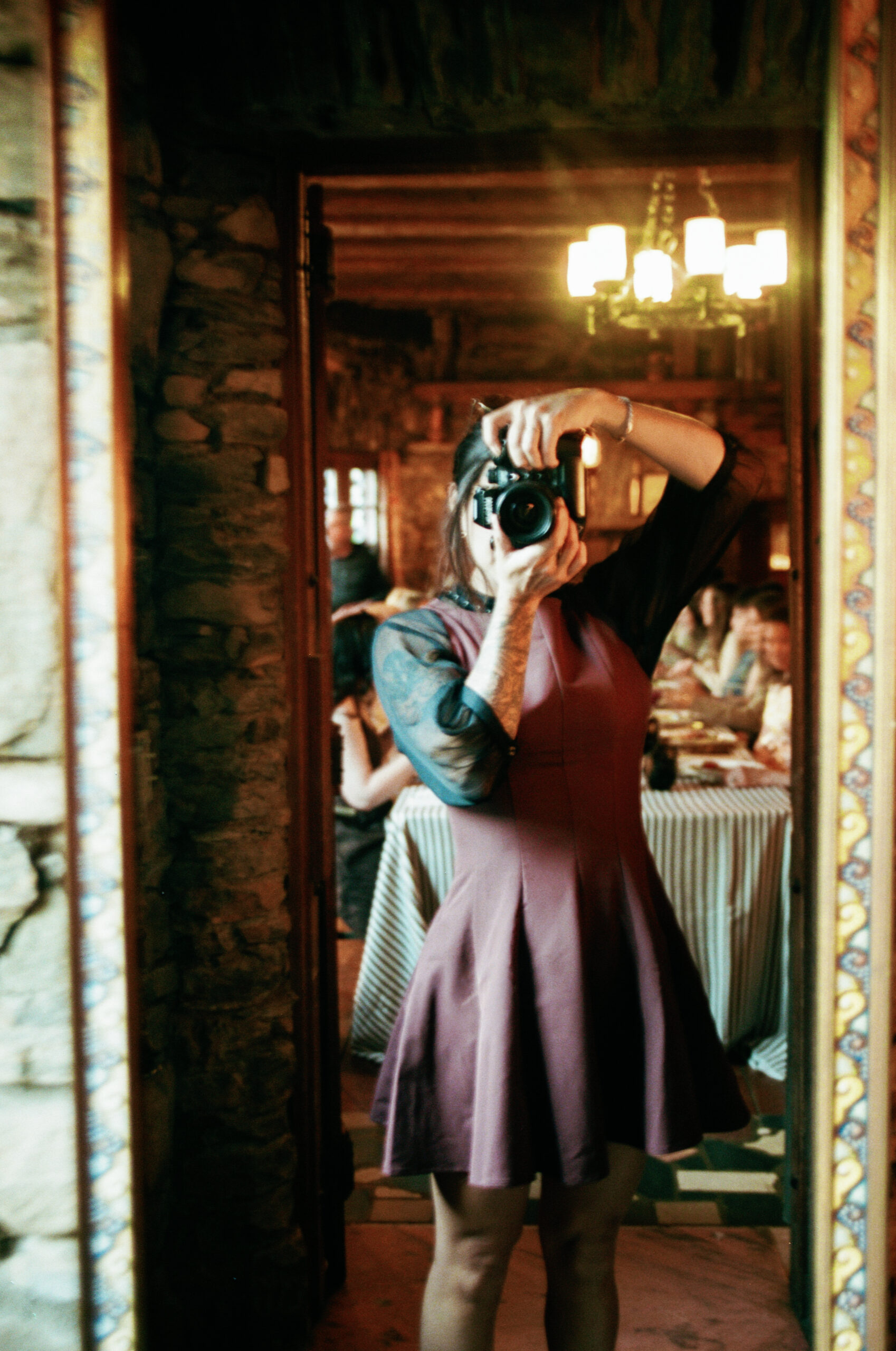 A person in a purple dress takes a mirror selfie inside a rustic, warmly lit room with stone walls—capturing a candid moment reminiscent of an Asheville Wedding Photographer's style, with people sitting at tables in the background. Asheville Douglas Ellington House wedding photographer