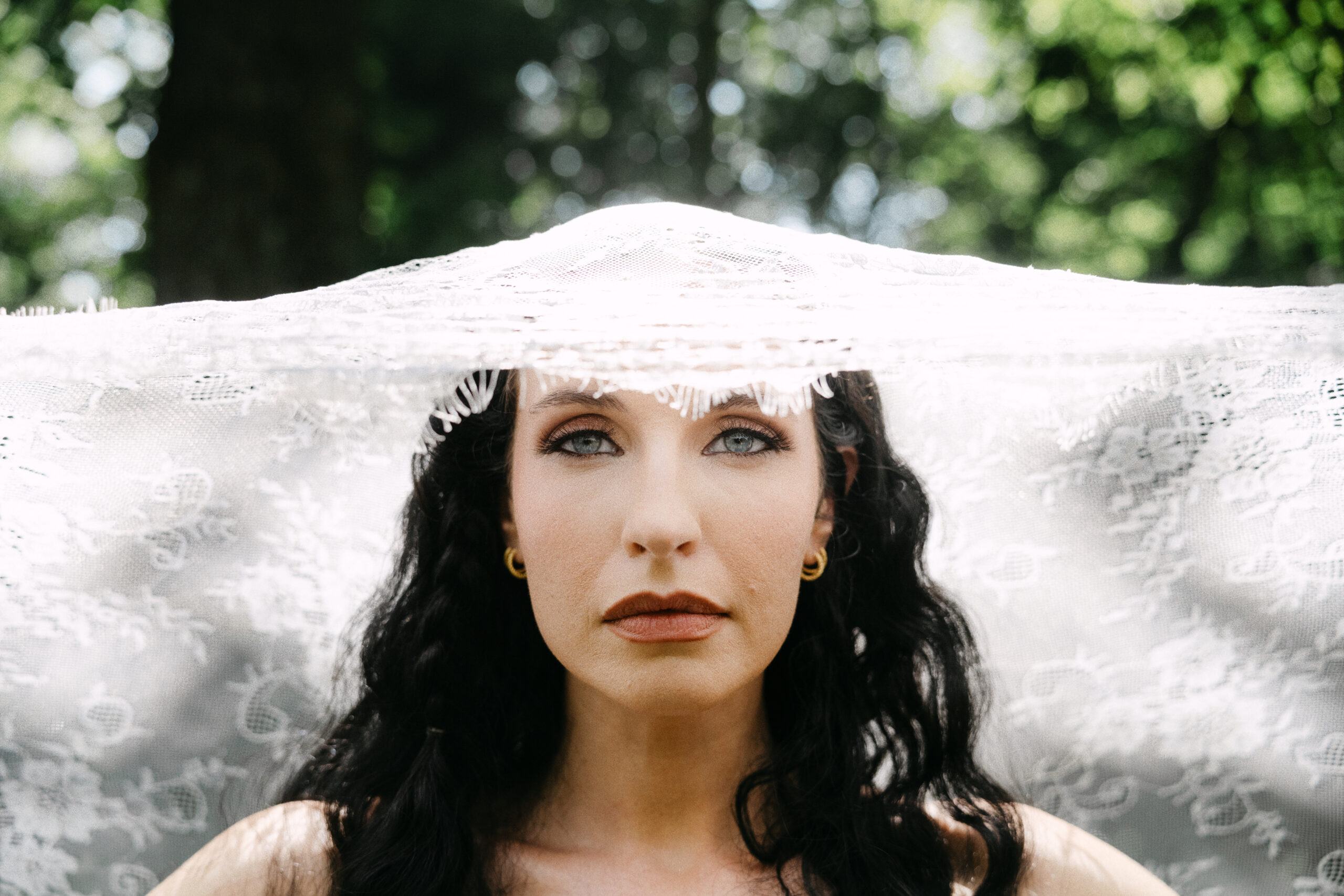 A woman with long dark hair and gold hoop earrings gazes forward, partially covered by a sheer lace veil, as an Asheville wedding photographer captures her serene expression against a backdrop of softly blurred greenery. Asheville wedding photographer
